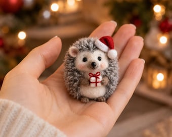 May include: A small needle-felted hedgehog figurine featuring grey and white fur, a tiny red and white Santa hat, and a miniature white gift box tied with a thin red ribbon held in its front paws. The hedgehog has a black nose and eyes.