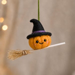 May include: A charming Halloween decoration. An orange pumpkin with a smiling face, wearing a black witch's hat with a purple band, is perched on a broom. The ornament is suspended by a green string, ideal for seasonal decor.