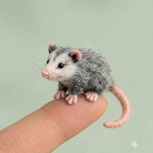 Puede incluir: Una figura miniatura de zarigüeya hecha de fieltro con un cuerpo gris difuso, cara blanca, orejas pequeñas rosadas, nariz rosa y una cola larga rosada. La pequeña criatura tiene ojos negros y bigotes finos con un acabado de textura lanosa.
