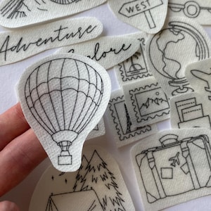 Op de afbeelding: Een set met zwart-witte stickers met een reisthema. De stickers bevatten afbeeldingen van een luchtballon, een koffer, een tent, een camera, een wereldbol, een vliegtuig, bergen, een busje en een bord met de tekst "Oost" en "West".