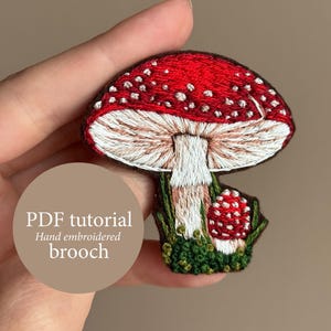 May include: Hand-embroidered mushroom brooch with a red cap dotted with white spots. The mushroom has a white stem and is surrounded by green grass. The text "PDF tutorial Hand embroidered brooch" is visible.