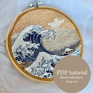 May include: Embroidered hoop art featuring a detailed depiction of Hokusai's "The Great Wave off Kanagawa." The artwork showcases a large wave in shades of blue and white, with a boat and Mount Fuji in the background. The text "PDF tutorial" is visible.