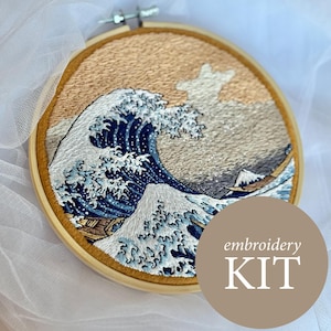 Könnte beinhalten: Stick-Set mit einer detaillierten Darstellung einer großen Meereswelle in Blau-, Weiß- und Goldtönen, die an das ikonische japanische Kunstwerk erinnert. Das Design befindet sich in einem hölzernen Stickrahmen. Der Text "embroidery KIT" ist sichtbar.