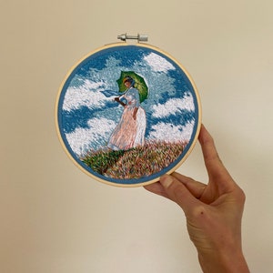 May include: Embroidered hoop art featuring a woman in a white dress holding a green umbrella, standing in a field of flowers under a blue sky with white clouds.