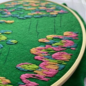 May include: An embroidery hoop with a green felt background featuring a colorful floral design. The design includes pink, orange, green, and blue embroidered flowers. The wooden hoop holds the fabric taut, showcasing the detailed needlework.