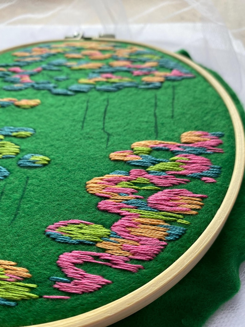 May include: An embroidery hoop with a green felt background featuring a colorful floral design. The design includes pink, orange, green, and blue embroidered flowers. The wooden hoop holds the fabric taut, showcasing the detailed needlework.