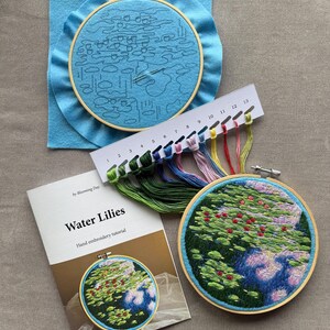 May include: An embroidery kit with a "Water Lilies" hand embroidery tutorial. The kit includes a blue felt hoop with a sketched design, a completed embroidery hoop, and a color chart with various thread colors. The finished embroidery depicts water lilies.