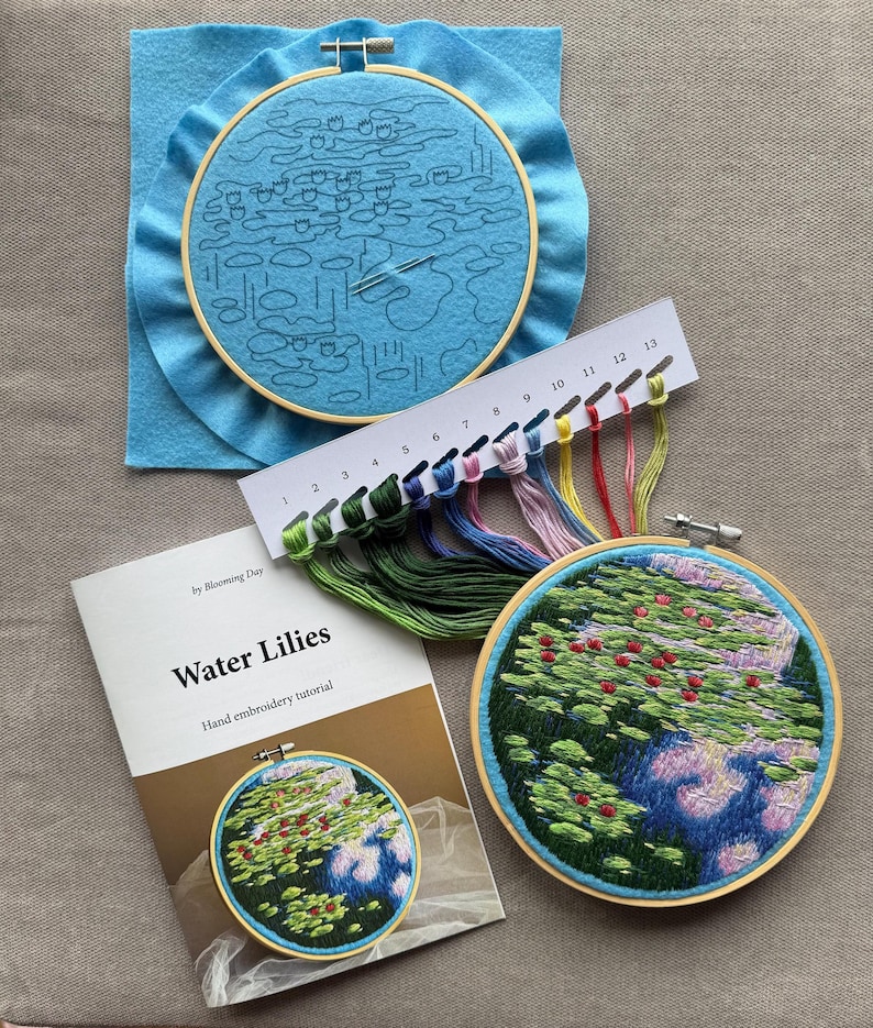 May include: An embroidery kit with a "Water Lilies" hand embroidery tutorial. The kit includes a blue felt hoop with a sketched design, a completed embroidery hoop, and a color chart with various thread colors. The finished embroidery depicts water lilies.