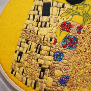 Pu&ograve; includere: Opera ricamata di Il Bacio di Gustav Klimt, che rappresenta una coppia che si abbraccia in un motivo a mosaico con accenti dorati, neri e rossi. L'opera &egrave; incorniciata in un cerchio di legno.