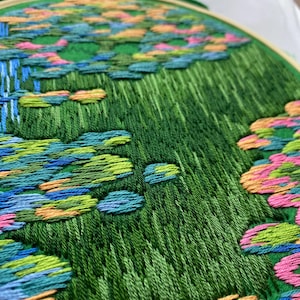 May include: Hand-stitched embroidery art in a wooden hoop. The design features a vibrant green field with colorful accents of blue, pink, and orange, resembling a landscape. The embroidery is detailed and textured, showcasing the craft's intricate nature.