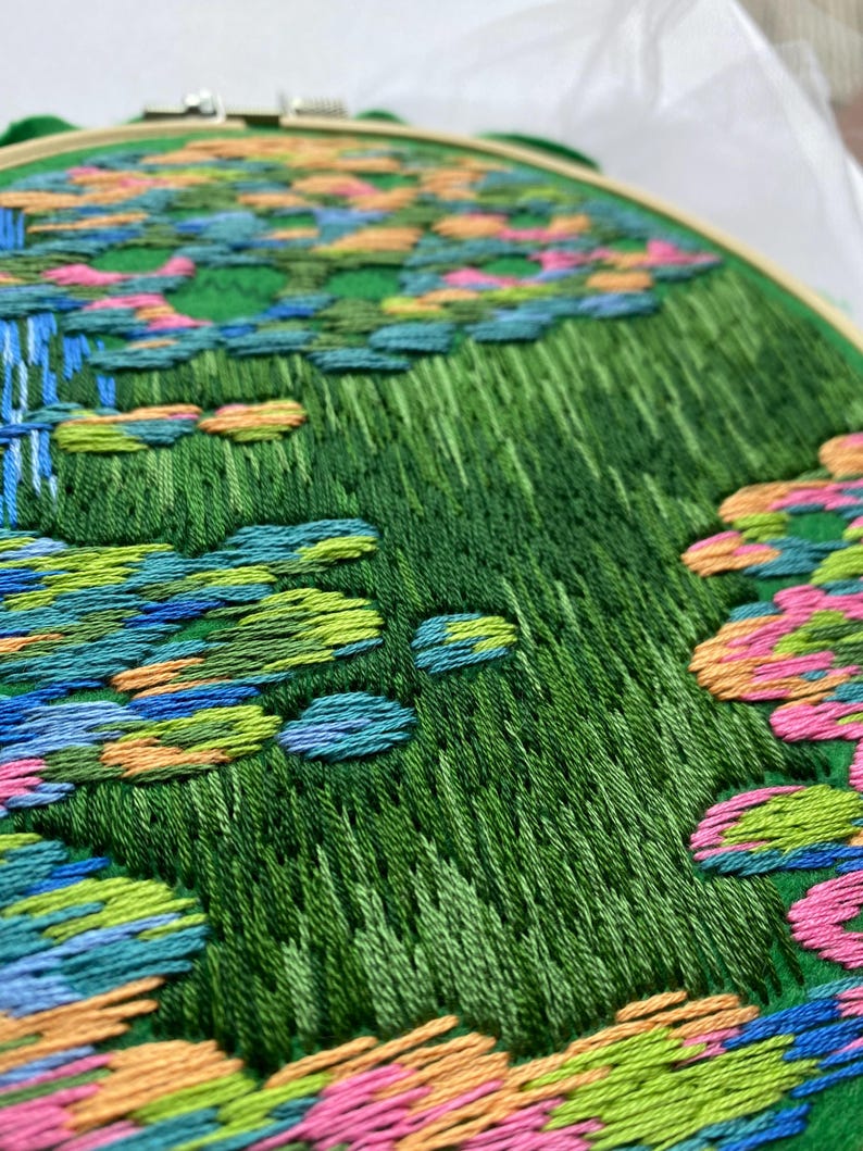 May include: Hand-stitched embroidery art in a wooden hoop. The design features a vibrant green field with colorful accents of blue, pink, and orange, resembling a landscape. The embroidery is detailed and textured, showcasing the craft's intricate nature.