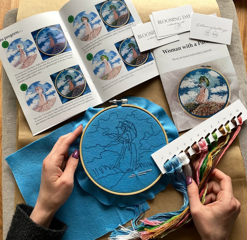 May include: A hand embroidery kit with a blue felt background, a wooden embroidery hoop with a blue fabric inside, and a set of embroidery floss in various colors. The kit includes instructions for a woman with a parasol embroidery project.