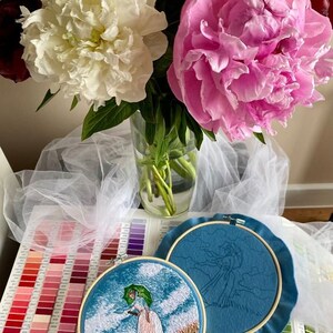 May include: Two embroidery hoops with blue fabric. One hoop shows a completed embroidery of a woman with an umbrella walking in a field. The other hoop shows an unfinished embroidery of a woman with an umbrella walking in a field.