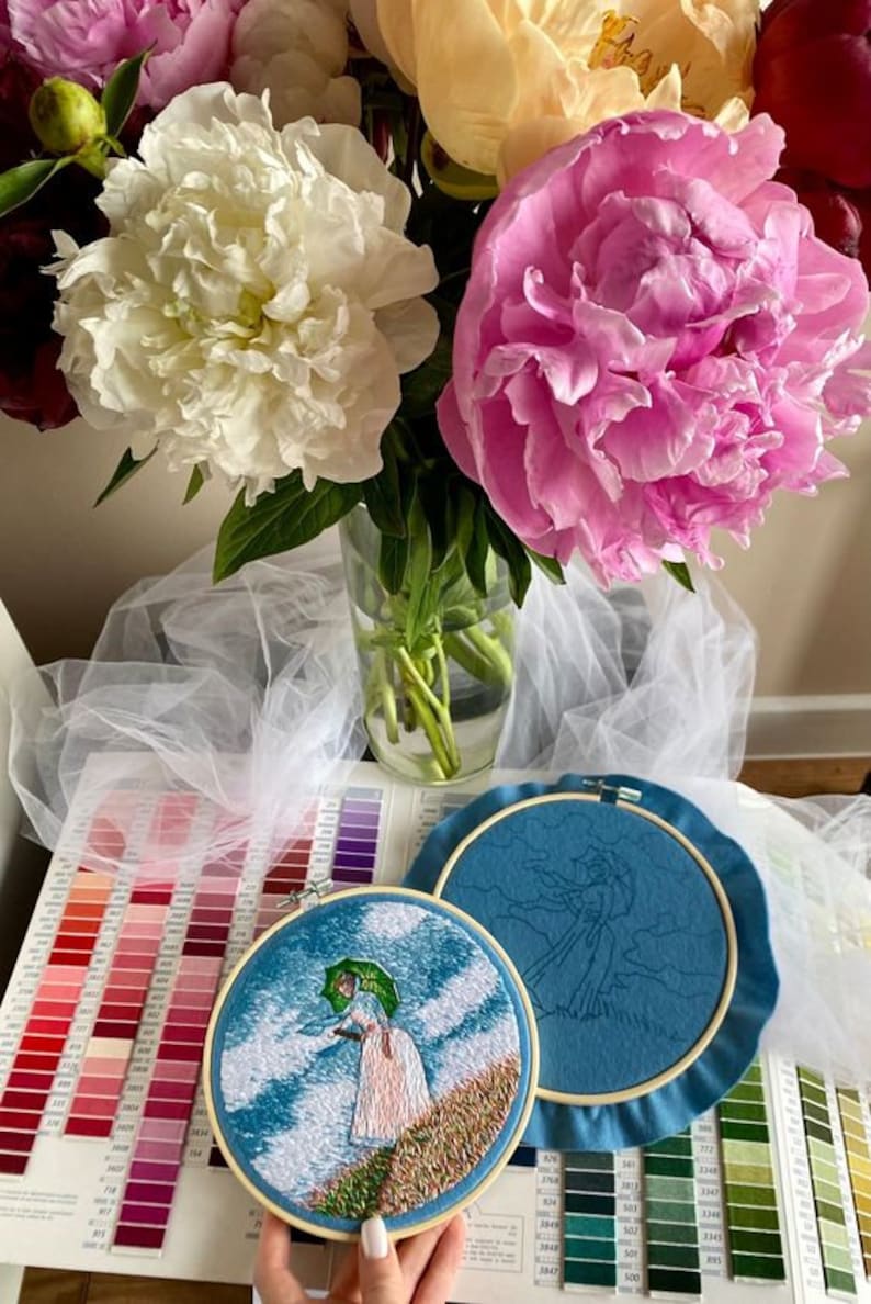 May include: Two embroidery hoops with blue fabric. One hoop shows a completed embroidery of a woman with an umbrella walking in a field. The other hoop shows an unfinished embroidery of a woman with an umbrella walking in a field.