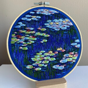 May include: Hand-stitched embroidery art of a water lily pond. The artwork is displayed in a round wooden hoop, featuring blue water, green lily pads, and pink and yellow flowers. The hoop is mounted on a small wooden stand.