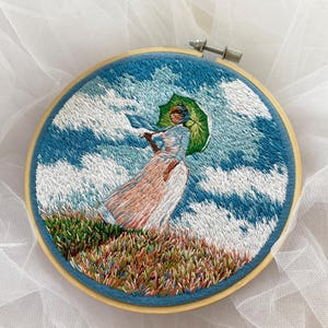 May include: Embroidered artwork in a wooden hoop depicts a person holding a green umbrella in a field of grass. The background features a blue sky with white clouds. The person is wearing a long dress.