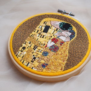 Pu&ograve; includere: Opera ricamata de Il Bacio di Gustav Klimt, che rappresenta una coppia che si abbraccia in un motivo a mosaico dorato. L'opera &egrave; incorniciata in un cerchio di legno con un retro in tessuto giallo.