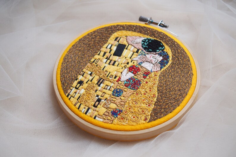 Pu&ograve; includere: Opera ricamata de Il Bacio di Gustav Klimt, che rappresenta una coppia che si abbraccia in un motivo a mosaico dorato. L'opera &egrave; incorniciata in un cerchio di legno con un retro in tessuto giallo.