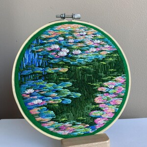 May include: Hand-stitched embroidery art depicting a serene water lily pond. The artwork, framed in a green hoop, features vibrant blues, greens, pinks, and white, creating a colorful and detailed scene of water lilies and their reflections.