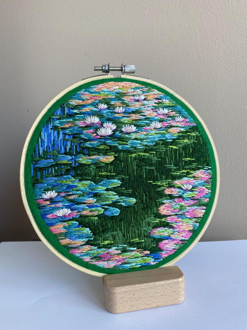 May include: Hand-stitched embroidery art depicting a serene water lily pond. The artwork, framed in a green hoop, features vibrant blues, greens, pinks, and white, creating a colorful and detailed scene of water lilies and their reflections.