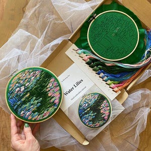 May include: An embroidery kit with the text "Water Lilies" includes a green fabric hoop with a floral design, embroidery floss, and a smaller finished piece. The kit is in a cardboard box with a white tulle fabric.