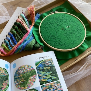 May include: Embroidery kit featuring a green fabric stretched in a wooden hoop, with a pre-printed design of a water garden. Includes colorful embroidery floss, needles, and a guide with the text "In progress...".
