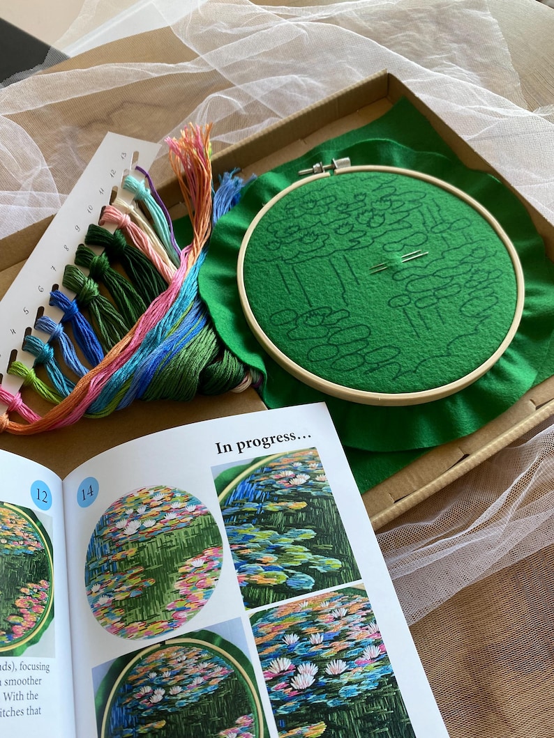 May include: Embroidery kit featuring a green fabric stretched in a wooden hoop, with a pre-printed design of a water garden. Includes colorful embroidery floss, needles, and a guide with the text "In progress...".