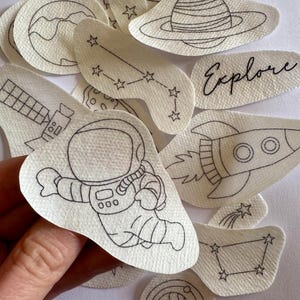 Op de afbeelding: Een set van 11 strijkbare patches met ruimtethema's, waaronder een astronaut, een raket, planeten, sterren en het woord "Explore".