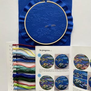 May include: A blue embroidery hoop with a partially stitched design, surrounded by blue fabric. A color chart with various thread colors and an instruction booklet are also present. The booklet reads "In progress..."