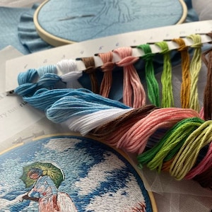 May include: A close-up of a cross-stitch embroidery project in progress. The embroidery hoop shows a woman with an umbrella standing in a field. A color chart with 10 different colored embroidery floss spools is shown in the foreground.