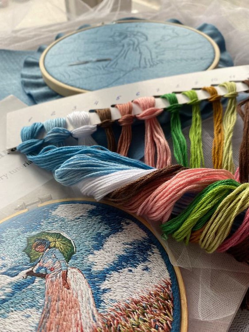 May include: A close-up of a cross-stitch embroidery project in progress. The embroidery hoop shows a woman with an umbrella standing in a field. A color chart with 10 different colored embroidery floss spools is shown in the foreground.