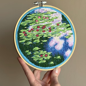 May include: Hand-stitched embroidery art in a wooden hoop. The artwork depicts a pond scene with green lily pads and vibrant red water lilies. The water is rendered in shades of blue and purple, creating a serene and detailed landscape.