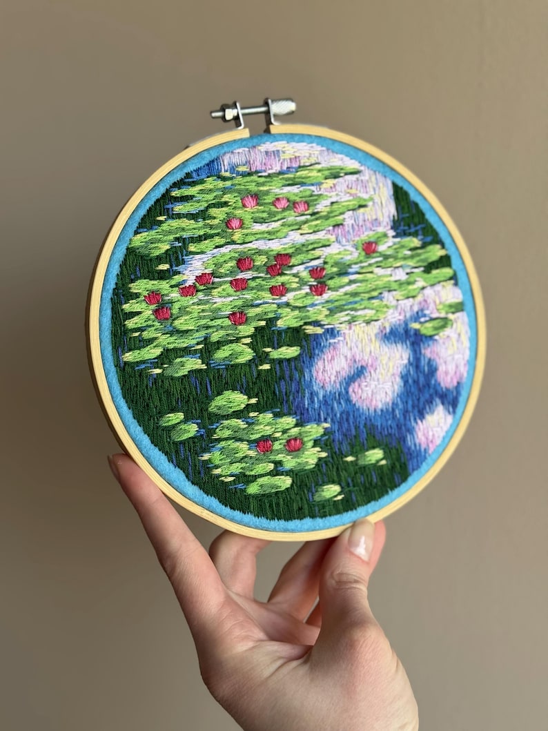 May include: Hand-stitched embroidery art in a wooden hoop. The artwork depicts a pond scene with green lily pads and vibrant red water lilies. The water is rendered in shades of blue and purple, creating a serene and detailed landscape.