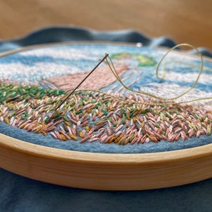 May include: Close-up of a partially completed embroidery hoop with a needle and thread. The embroidery features a landscape scene with green, pink, and brown stitches.
