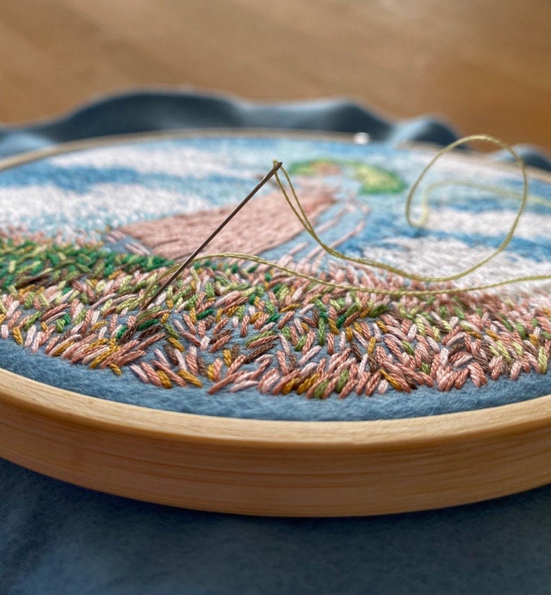 May include: Close-up of a partially completed embroidery hoop with a needle and thread. The embroidery features a landscape scene with green, pink, and brown stitches.