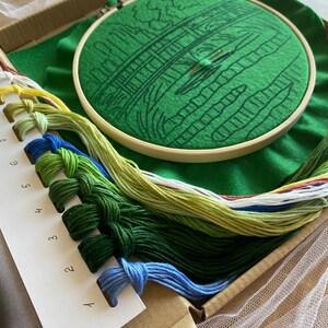 May include: Embroidery kit featuring a green felt hoop with a bridge design, a needle, and various colored embroidery floss. The kit includes a cardboard box, a green felt background, and a piece of white tulle.