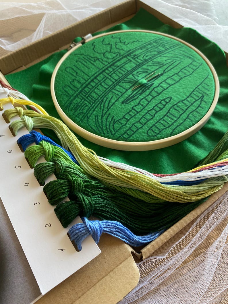 May include: Embroidery kit featuring a green felt hoop with a bridge design, a needle, and various colored embroidery floss. The kit includes a cardboard box, a green felt background, and a piece of white tulle.