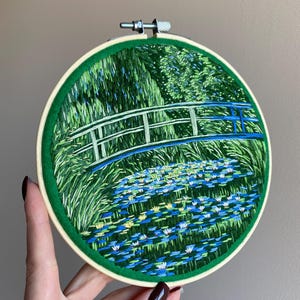 May include: Hand-stitched embroidery art in a round hoop. The design features a bridge over a pond filled with lily pads, with green foliage in the background. The bridge has blue and white accents, and the hoop is trimmed with green fabric.