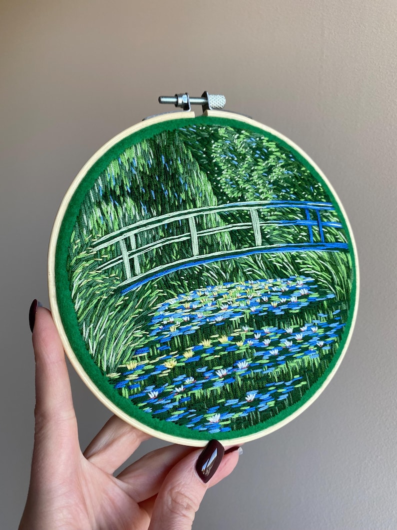 May include: Hand-stitched embroidery art in a round hoop. The design features a bridge over a pond filled with lily pads, with green foliage in the background. The bridge has blue and white accents, and the hoop is trimmed with green fabric.