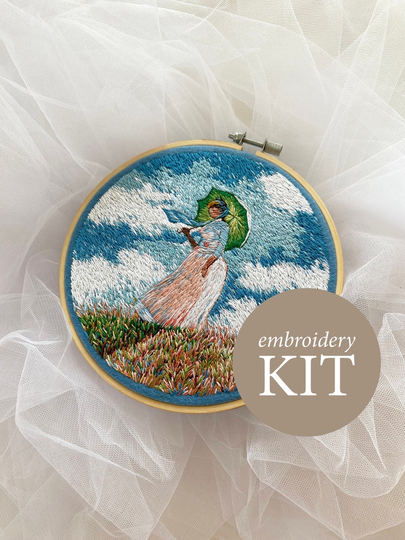 May include: Embroidery kit featuring a woman with an umbrella standing in a field with a blue sky and white clouds. The kit includes all the materials needed to complete the embroidery project. The text "embroidery KIT" is visible in the image.