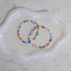 May include: Two colourful beaded bracelets with blue evil eye charms and yellow smiley face charms.