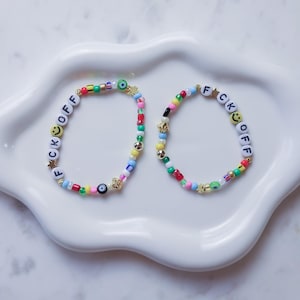 May include: Two colourful beaded bracelets with the phrase "F*CK OFF" spelled out in white beads. The bracelets are on a white ceramic dish.
