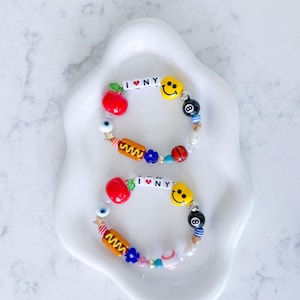 May include: Two elastic beaded bracelets featuring white letter beads spelling I heart NY, red apple charms, yellow smiley faces, black eight-ball beads, hot dog charms, blue flower beads, evil eye beads, and various colourful accent beads.