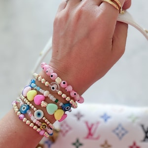 以下が含まれることがあります： ピンク、青、黄色、金など、さまざまな色のビーズブレスレットのコレクション。一部のブレスレットには、ハートと邪視のチャームが付いています。ブレスレットは手首に重ねられ、指には指輪があります。カラフルな模様の白いハンドバッグが背景にあります。