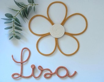 May include: A wall decoration featuring a five-petaled flower made of mustard yellow rope with a white coiled rope center, accompanied by the name Lisa written in cursive using coral-colored rope and a small green eucalyptus sprig.