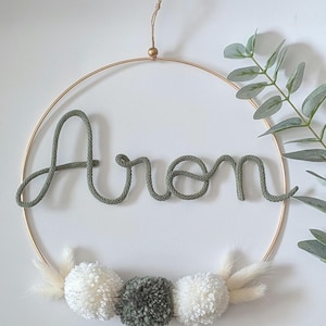 Peut inclure: Un cercle doré avec le nom "Aron" en corde verte. Le bas est orné de pompons blancs et gris et de fleurs séchées. Un élément décoratif pour une chambre d'enfant.