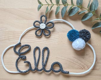 May include: A white rope cloud with the name "Ella" spelled out in black rope. The cloud is decorated with a black and white flower and three pom-poms in blue, gray, and white.