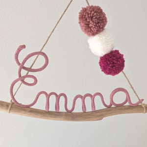 May include: A pink rope name "Emma" is displayed on a piece of driftwood, with three pom-poms in shades of pink and white hanging above. The name is attached to the wood with twine, creating a decorative wall hanging.