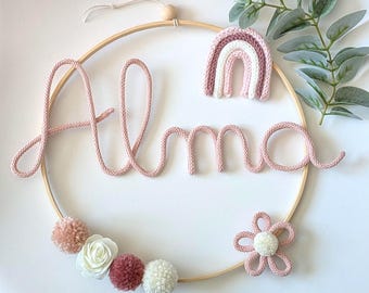 May include: A circular wall hanging with the name "Alma" in pink yarn. The design includes a knitted rainbow, a flower, pom-poms, and faux greenery. The wooden hoop is adorned with decorative elements, creating a charming nursery or room decor.