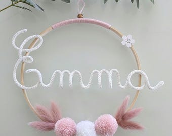 May include: A circular wall hanging with the name "Emma" crafted in white yarn. The wooden hoop is adorned with pink yarn, pom-poms, and dried flowers. A small white flower is attached to the hoop.